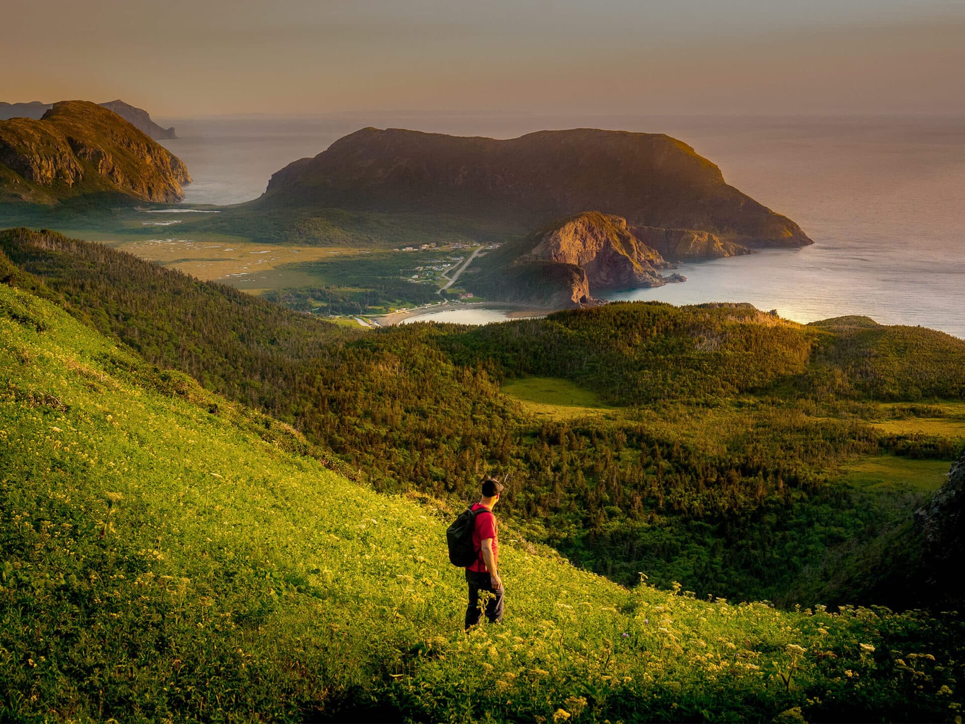 A person with a backpack stands on a green hillside overlooking a scenic coastal landscape with mountains, cliffs, and the sea at sunset.